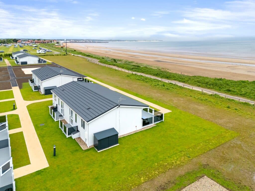 Save the dates for our upcoming events at South Shore Park 6 Aerial view of a beach with several holiday chalets nestled along the shoreline, surrounded by greenery and blue water.