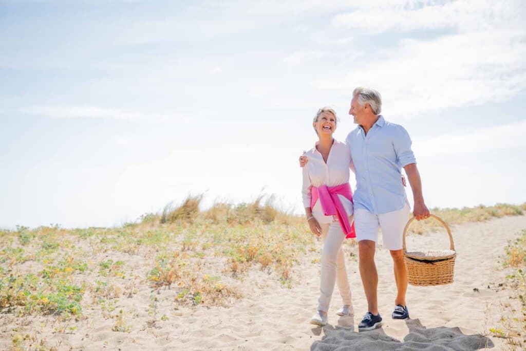 Save the dates for our upcoming events at South Shore Park 4 An elderly couple walks hand in hand on the beach, a picnic basket in tow, surrounded by sand and waves.